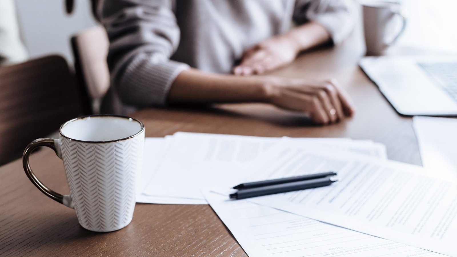 Hände auf einem Schreibtisch mit Abrechnungsunterlagen, Stift, Kaffeetasse und Laptop